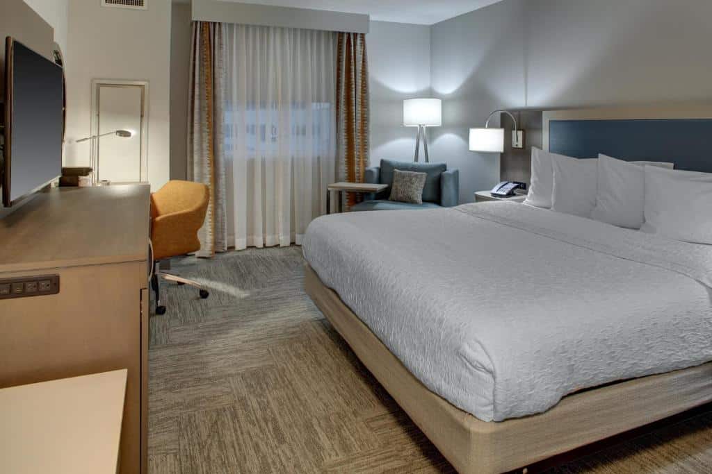 Quarto no Hampton Inn & Suites Miami Airport South/Blue Lagoon com cama king size, escrivaninha e cadeira, poltrona com abajur, janela grande com cortinas e decoração neutra, para representar hotéis com cozinha em Miami.