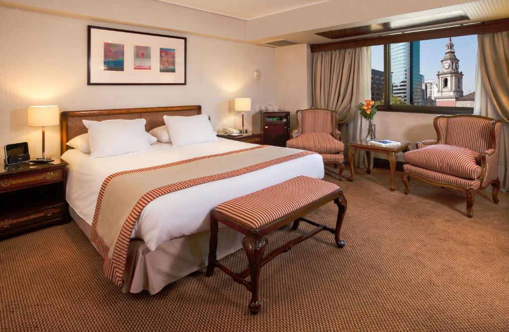 Quarto do Hotel Plaza San Francisco com cama de casal do lado esquerdo, duas poltronas ao lado de madeira com luminária do lado esquerdo do quarto e duas poltronas estofadas.