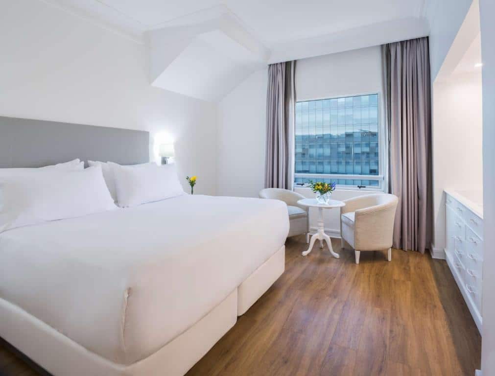 Quarto do NH Collection Plaza com cama de casal no centro do quarto do lado esquerdo do quarto duas poltronas brancas com um mesa redonda no meio.