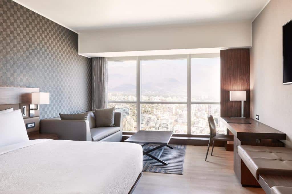 Quarto do AC Hotel by Marriott Santiago Costanera Center com cama de casal do lado esquerdo ao lado da cama, um sofá com dois lugares com uma mesa no centro e em frente a mesa uma mesa de trabalho com cadeira.