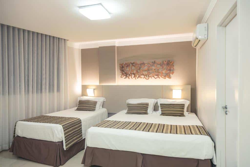 Quarto do Lagoon Prime Hotel, com uma cama de casal e uma de solteiro estão lado a lado, uma delas ao lado da porta e outra ao lado da janela, é possível ver também duas luminárias e um ar-condicionado, para representar hotéis e pousadas em Lagoa Santa.