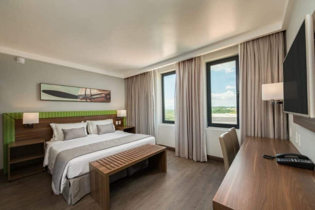 Quarto do hotel Linx Confins, com uma cama de casal e uma banqueta em sua ponta estão no meio, do lado de duas janelas e cortinas, em frente a cama está uma mesa com cadeira e uma tv pendurada na parede, para representar hotéis e pousadas em Lagoa Santa.