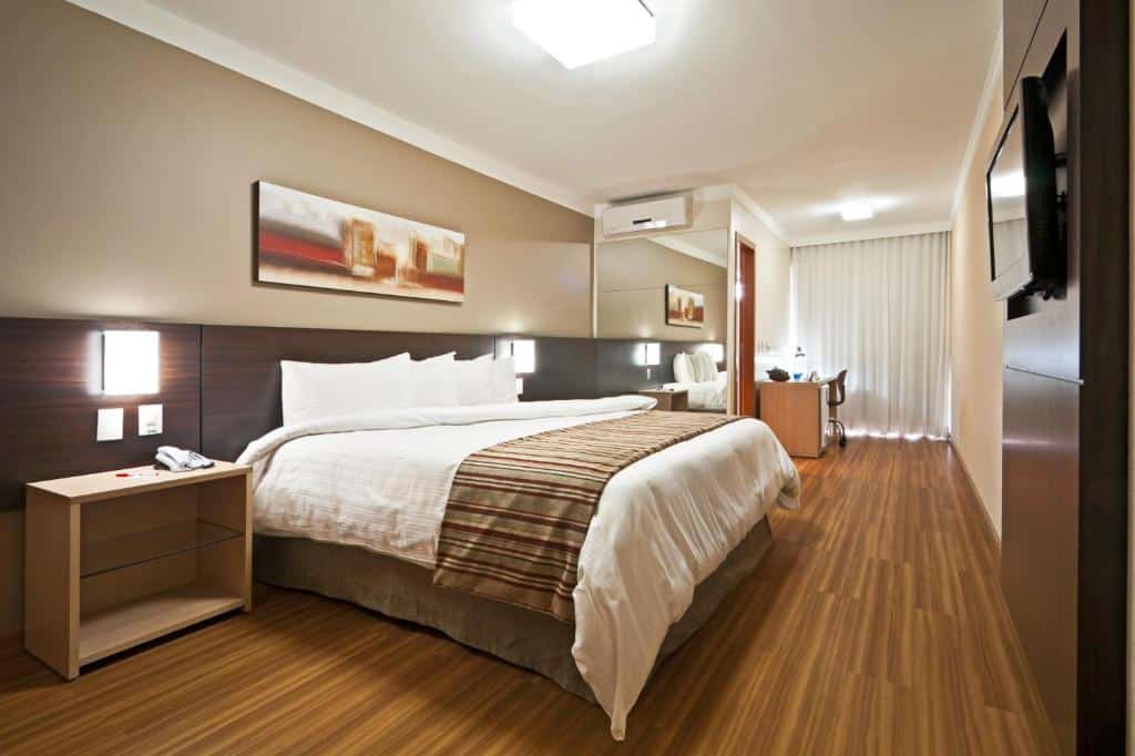 Quarto do Ramada Hotel & Suites Lagoa Santa By Wyndham, com uma cama de casal está no meio, ao lado de uma mesinha. Ao fundo, na parte direita, há um espelho, uma mesa maior com cadeira e cortinas cobrindo a janela, para representar hotéis e pousadas em Lagoa Santa.