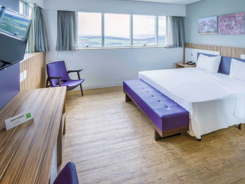 Quarto do ibis Styles Confins Aeroporto, com uma cama de casal no meio do quarto, em frente há uma mesa embutida na parede e uma tv, ao fundo está uma janela e uma cadeira, para representar hotéis e pousadas em Lagoa Santa.