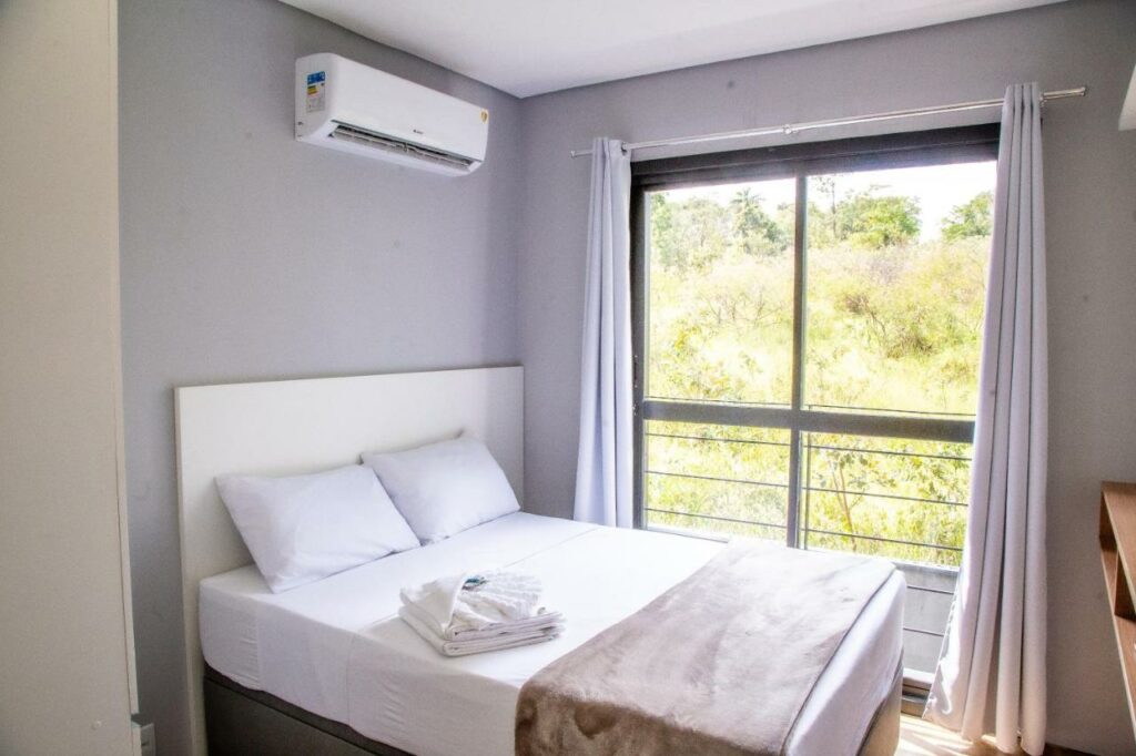 Quarto do Mini House Lagoa Santa, com uma cama de casal estão ao lado de uma porta que leva até a varanda, mostrando várias árvores do lado de fora, para representar hotéis e pousadas em Lagoa Santa.