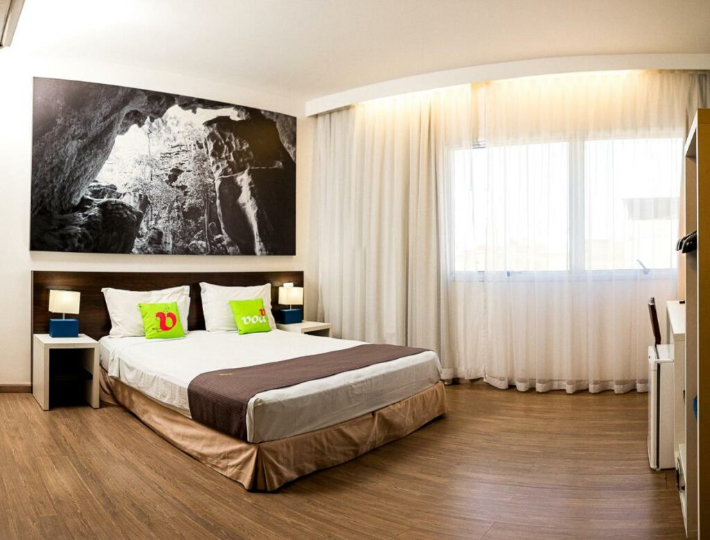 Quarto do VOA Business Supreme Choice Confins, com uma cama de casal está bem no meio, entre duas luminárias, ao seu lado há uma janela coberta por grandes cortinas, para representar hotéis e pousadas em Lagoa Santa.