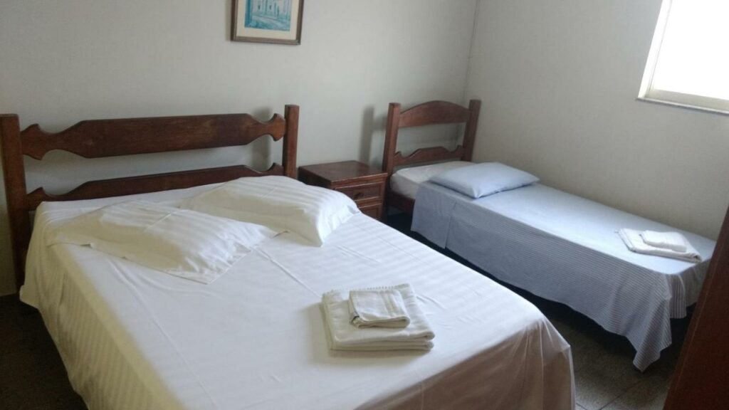 Quarto do Hotel Lund, com uma cama de casal e uma cama de solteiro estão lado a lado, entre elas há uma mesinha pequena, para representar hotéis e pousadas em Lagoa Santa.