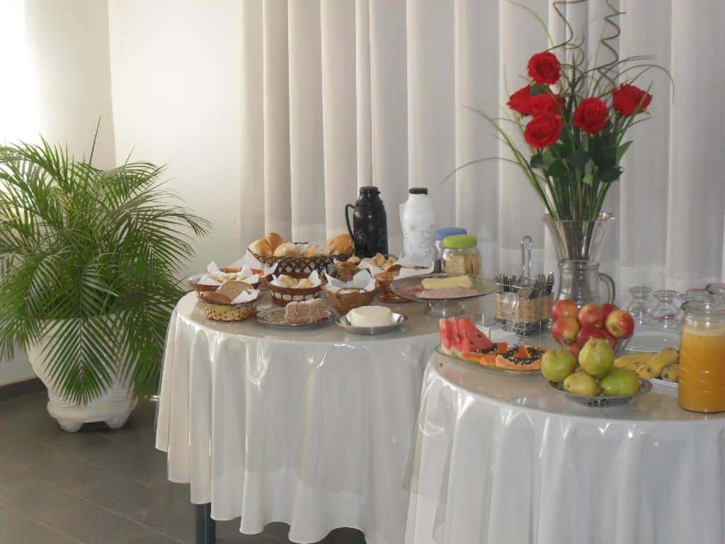 Mesas de café da manhã do Hotel Lund, são duas mesas redondas com frutas, frios, pães, queiro e algumas outras coisas. Um vaso de flor decorativo também está ali.