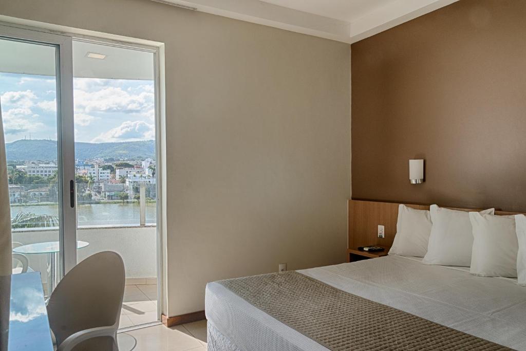 Quarto do Hotel Atlas, que representa o post sobre hotéis e pousadas em Sete Lagoas. Na direita vemos a cama box de casal, e ao seu lado esquerdo há uma mesa de cabeceira com tomada e controles em cima. Atrás da cama, há uma parede marrom com um pequeno abajur fixo. No canto inferior esquerdo da foto, na frente da cama, vemos o relance de uma escrivaninha com uma cadeira. Do lado dessa mesa, ao fundo do quarto, há uma porta branca de correr que dá acesso para a varanda.
