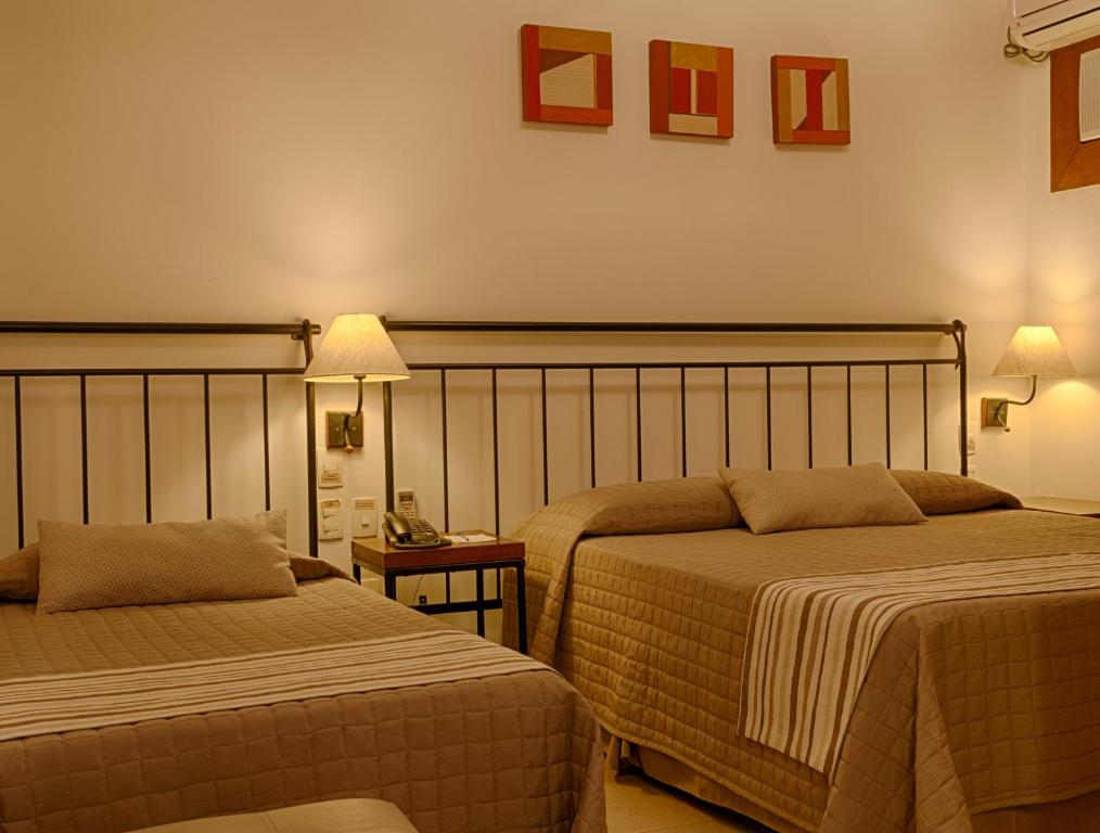 Quarto do Hotel Vila Serrana, que representa o post sobre hotéis e pousadas em Sete Lagoas. Vemos duas camas, uma de casal na direita e outra de solteiro na esquerda. No meio delas, há uma pequena mesa de cabeceira com um telefone preto em cima, e atrás, um abajur, algumas tomadas e interruptores. No canto superior direito, vemos o relance de um ar-condicionado.