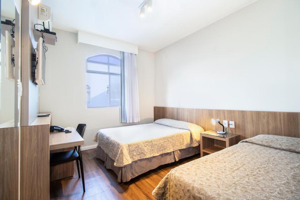 Quarto do Real Hotel, que representa o post sobre hotéis e pousadas em Sete Lagoas. Há duas camas no quarto, uma de solteiro no canto inferior esquerdo e outra ao seu lado, de casal. Entre elas há uma mesa de cabeceira com abajur em cima e uma tomada atrás. Há uma janela no fundo do quarto, do lado da cama de casal. E na frente dessa cama, há uma escrivaninha com cadeira, telefone e TV. 