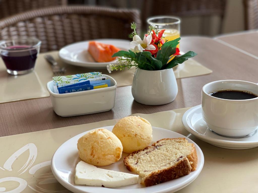 Imagem de um café da manhã no Real hotel. No prato que está no meio da foto, há dois pães de queijo, uma fatia de bolo e outra de queijo branco. Do lado direito, há uma xícara de café. No meio da mesa há um vaso de porcelana com flores e outro com sachês de açúcar e sal. Do outro lado da mesa há outro prato com uma fruta e dois copos com sucos.
