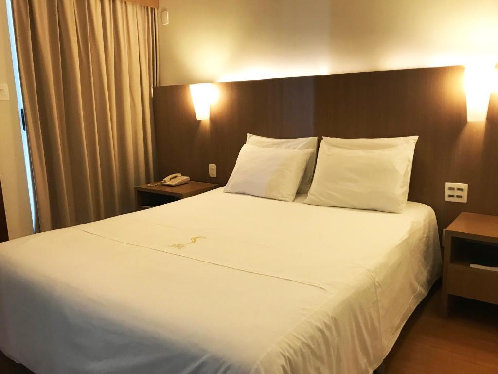 Quarto do Sete Lagoas Residence Hotel, que representa o post sobre hotéis e pousadas em Sete Lagoas. No centro há uma cama box de casal, e nos seus dois lados há uma mesa de cabeceira de madeira. Na mesa à esquerda, há um telefone em cima. Há dois abajures fixos na cabeceira da cama. Na esquerda, está a janela coberta por cortinas.