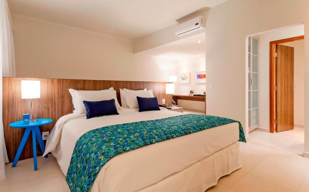 Quarto no Tulip Inn Sete Lagoas, que representa o post sobre hotéis e pousadas em Sete Lagoas. No meio há uma cama box de casal. Do seu lado esquerdo, há uma mesa azul pequena com abajur em cima. Do outro lado, uma mesa de cabeceira quadrada com gavetas e outro abajur. Seguindo para o fundo do quarto, do lado direito, vemos uma mesa de madeira com amenidades em cima, e também um ar-condicionado no topo da parede. A porta está no lado direito. 