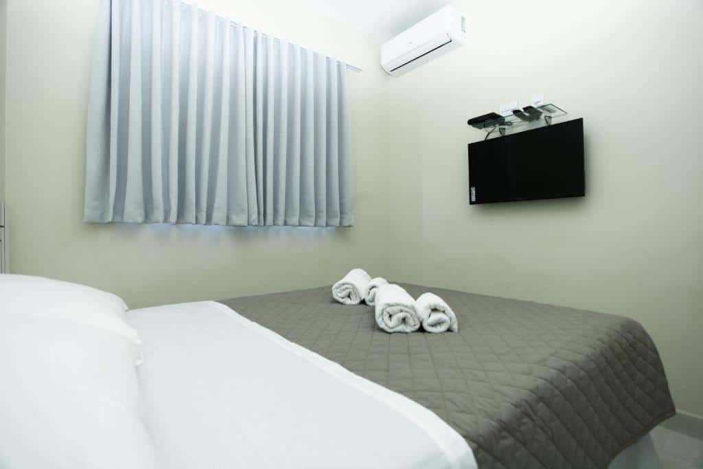 Quarto no Rivello Apart Hotel, que representa o post sobre hotéis e pousadas em Sete Lagoas. Vemos a cama no ângulo de quem está deitado.. Há toalhas em cima e uma TV fixa na parede em sua frente. Na sua esquerda, há uma janela coberta por cortinas cinzas.
