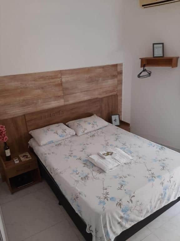 Foto do quarto da kitnet apart hotel 3 que representa o post sobre hotéis e pousadas em Sete Lagoas. No centro, vemos uma cama box de casal com tolhas em cima. Possui uma mesa de cabeceira no lado direito e esquerdo com alguns itens decorativo em cima.
