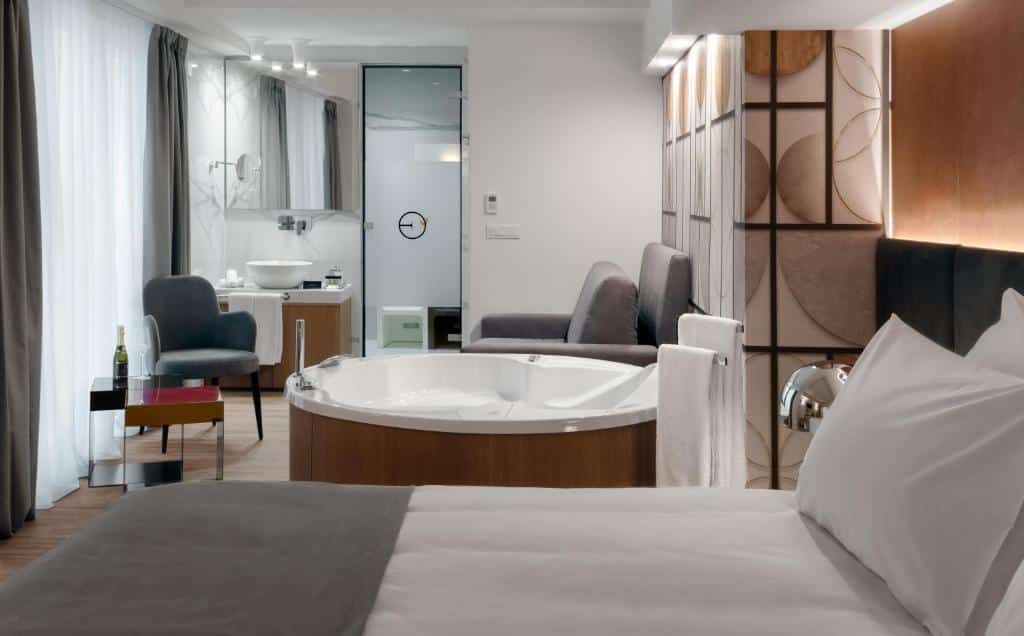 Quarto moderno no Elia Ermou Athens Hotel com banheira redonda, cama, área de estar e banheiro com paredes de vidro em um esquema de cores neutras, para representar hotéis em Atenas.