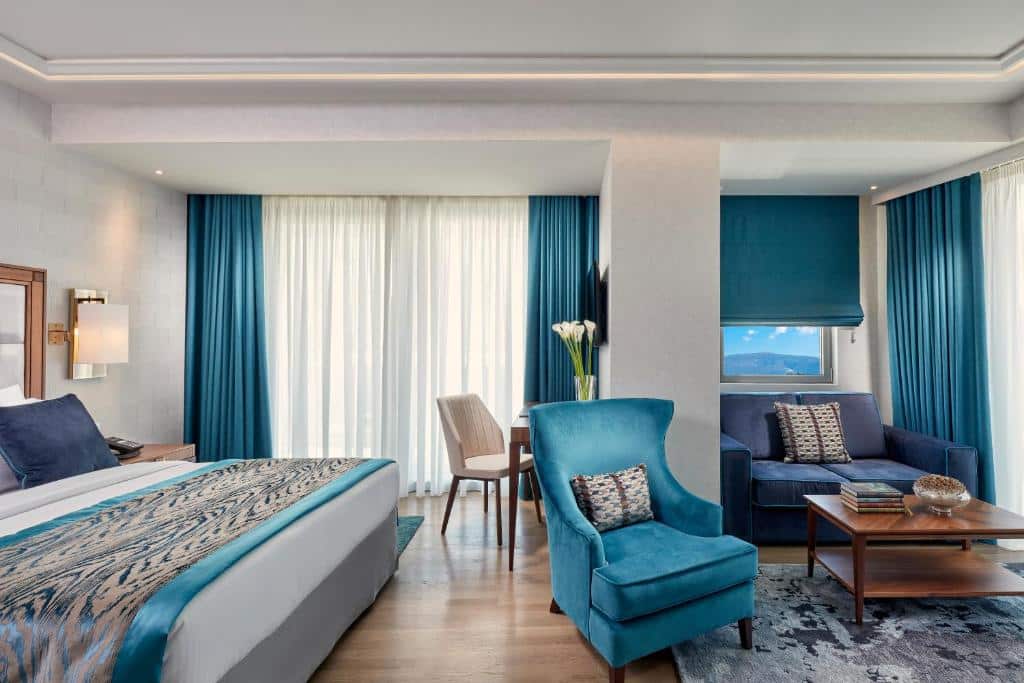 Quarto moderno no Electra Metropolis com uma cama grande, poltrona azul-petróleo, sofá, escrivaninha, piso de madeira e janelas com cortinas azuis, para representar hotéis em Atenas.