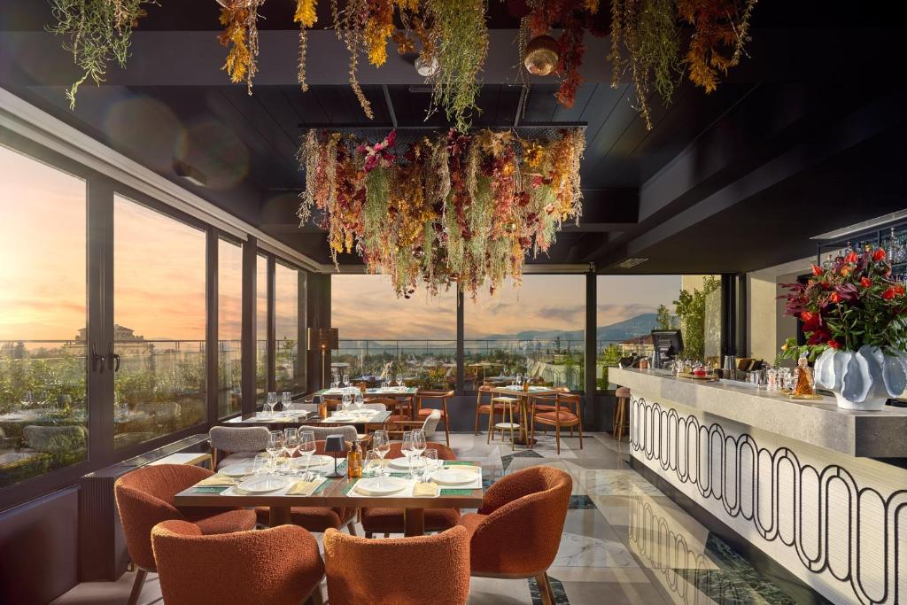 Restaurante moderno com grandes janelas, cadeiras laranja, mesas postas e flores secas penduradas, com vista para o pôr do sol, na propriedade do Stay Hotel.