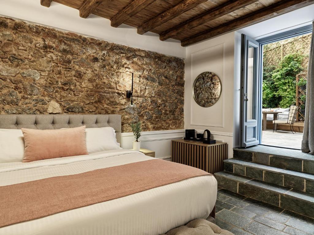 Quarto aconchegante no A77 Suites by Andronis com parede com detalhes em pedra, cama de casal, espelho redondo, porta aberta para o pátio e vigas de madeira natural no teto, para representar hotéis em Atenas.