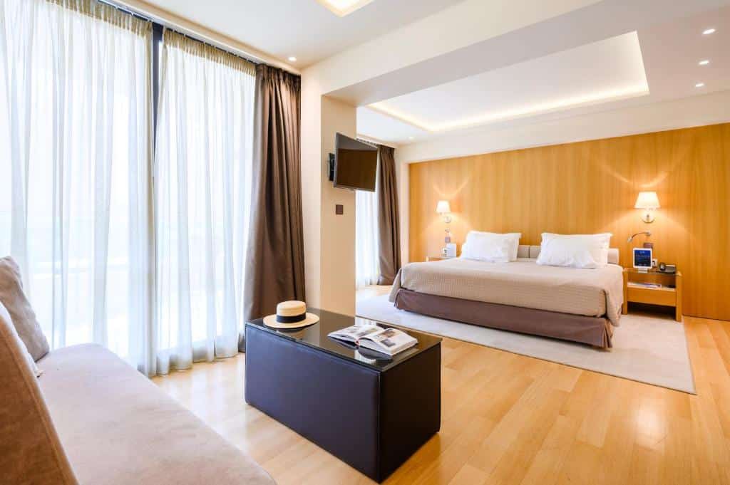 Quarto moderno no Somewhere Vouliagmeni com uma cama grande, sofá, TV na parede, piso de madeira e cortinas do chão ao teto, para representar hotéis em Atenas.