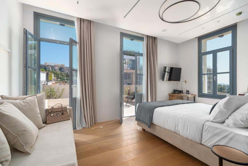 Quarto moderno no Hellenic Vibes Smart Hotel com janelas grandes, uma cama, sofá, TV na parede, escrivaninha pequena e piso de madeira, para representar hotéis em Atenas.