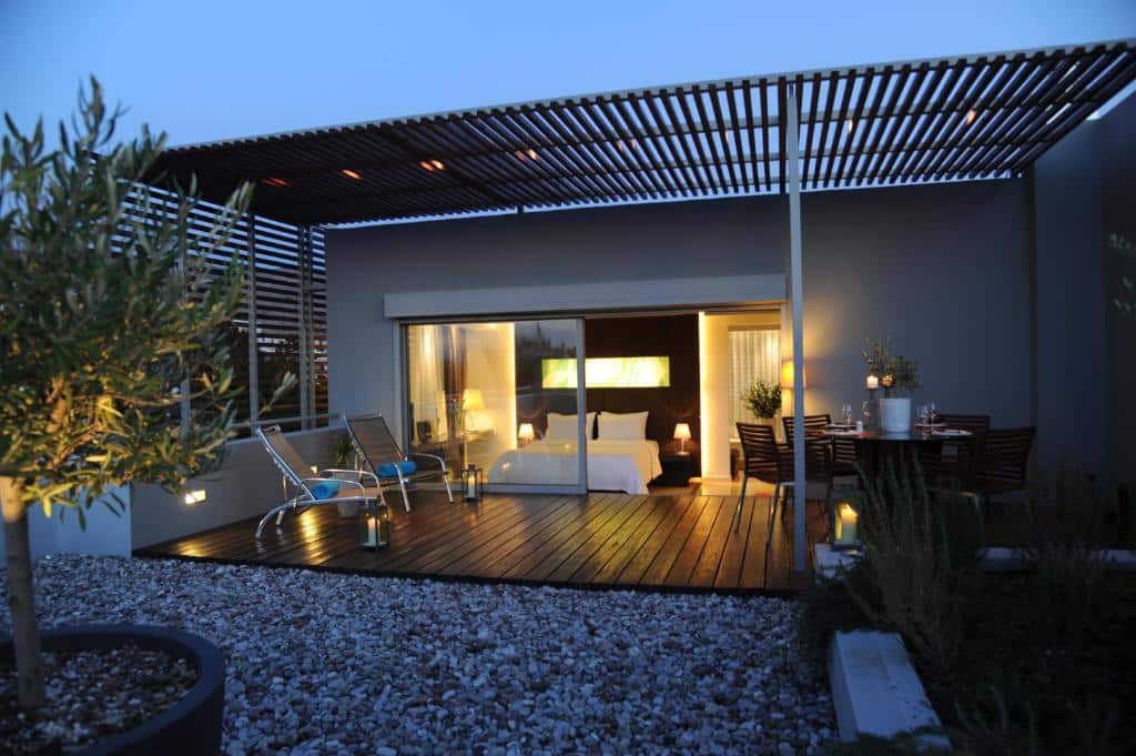 Pátio moderno no Brasil Suites Boutique Hotel ao anoitecer com espreguiçadeiras, mesa de jantar, deck de madeira, cascalho e grandes portas de vidro que levam a um quarto iluminado, para representar hotéis em Atenas.