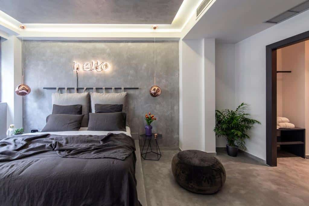 Quarto moderno no 14 Reasons Why Hotel Athens com paredes e roupa de cama cinza, letreiro "hello" em neon, luzes penduradas, plantas e um armário aberto, para representar hotéis em Atenas.