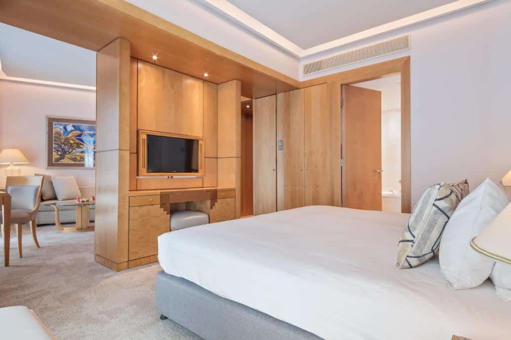 Quarto moderno no Melia Athens com uma cama grande, TV na parede, móveis de madeira e uma área de estar adjacente, para representar hotéis em Atenas.