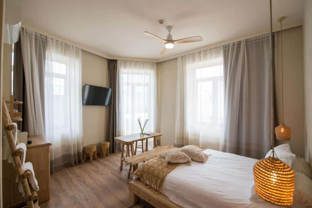 Um quarto bem iluminado no Ederlezi Boutique Hotel com uma cama, mesa pequena, cadeiras, TV na parede, piso de madeira e janelas grandes com cortinas transparentes, para representar hotéis em Atenas.