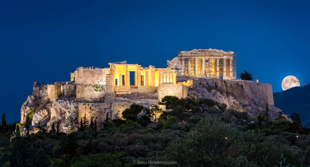 A Acrópole de Atenas é iluminada à noite com a lua cheia surgindo no céu azul claro ao fundo.