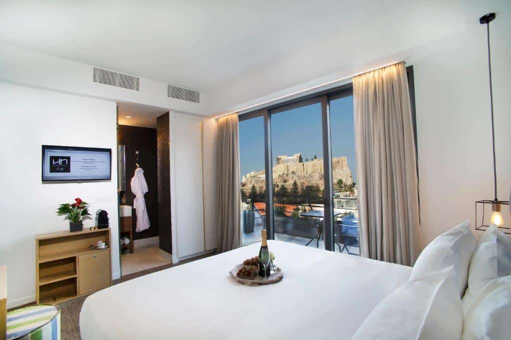 Quarto moderno no Nlh Fix | Neighborhood Lifestyle Hotels com uma cama grande, uma bandeja com vinho e frutas e vista para uma ruína antiga através de janelas do chão ao teto, para representar hotéis em Atenas.
