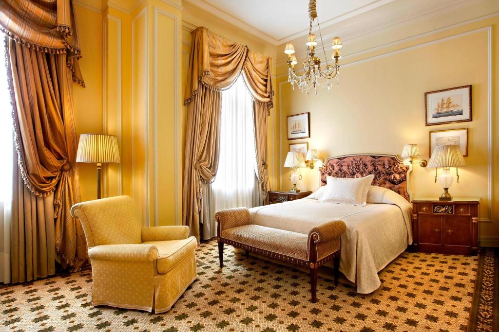 Um quarto clássico no Hotel Grande Bretagne, a Luxury Collection Hotel, Athens com uma cama de casal, poltrona, cortinas ornamentadas, lustre e duas mesas de cabeceira com luminárias, para representar hotéis em Atenas.