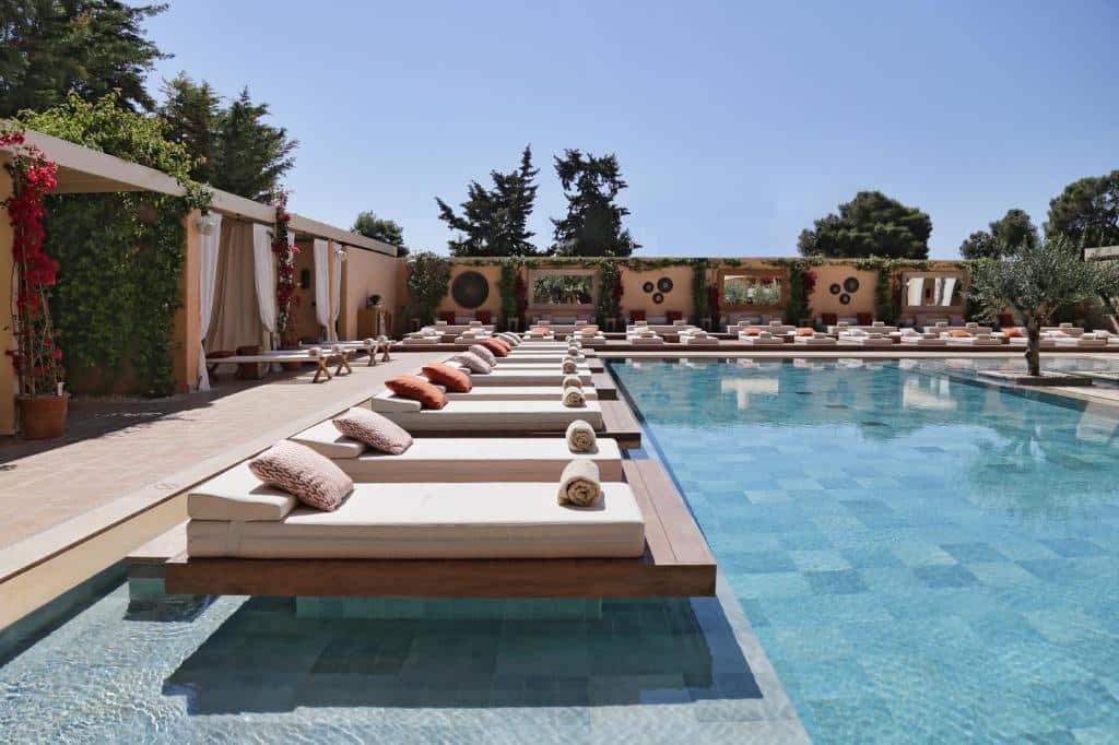 Espreguiçadeiras com almofadas e toalhas enroladas estão dispostas ao lado de uma piscina externa transparente em um ambiente ensolarado, na propriedade do The Margi.