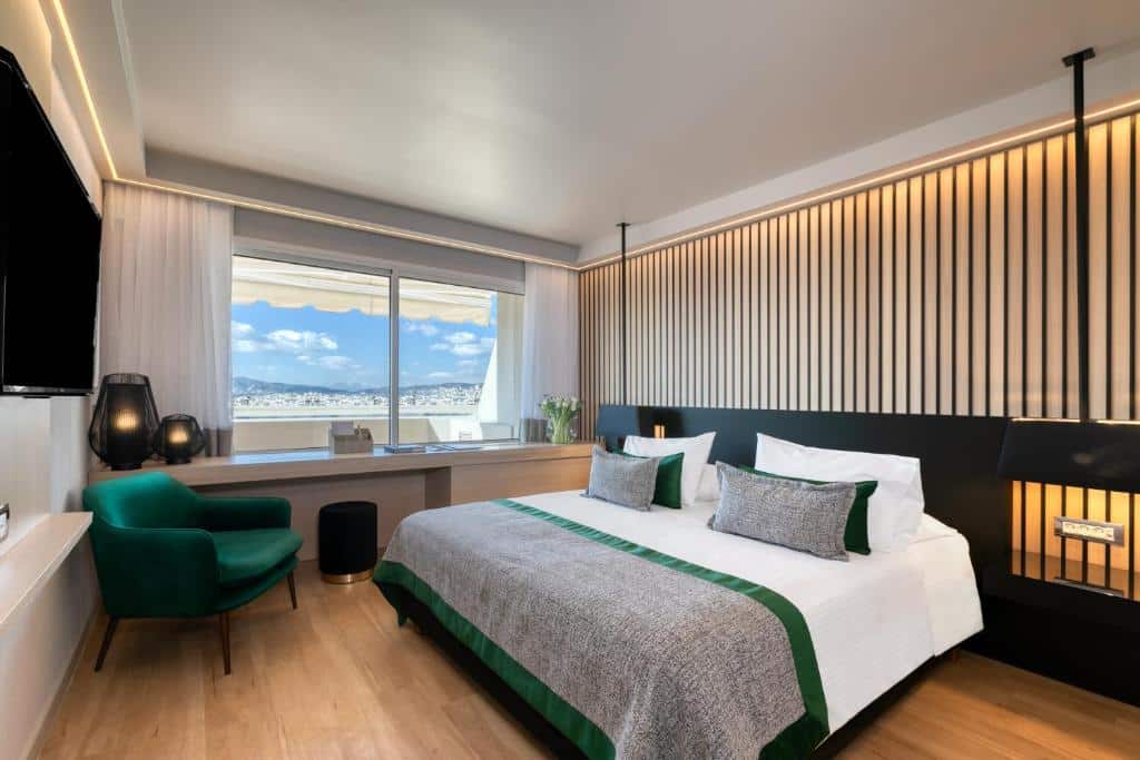 Quarto moderno no Divani Caravel com uma cama grande, poltrona verde, escrivaninha e uma janela com vista para o mar e o céu, para representar hotéis em Atenas.