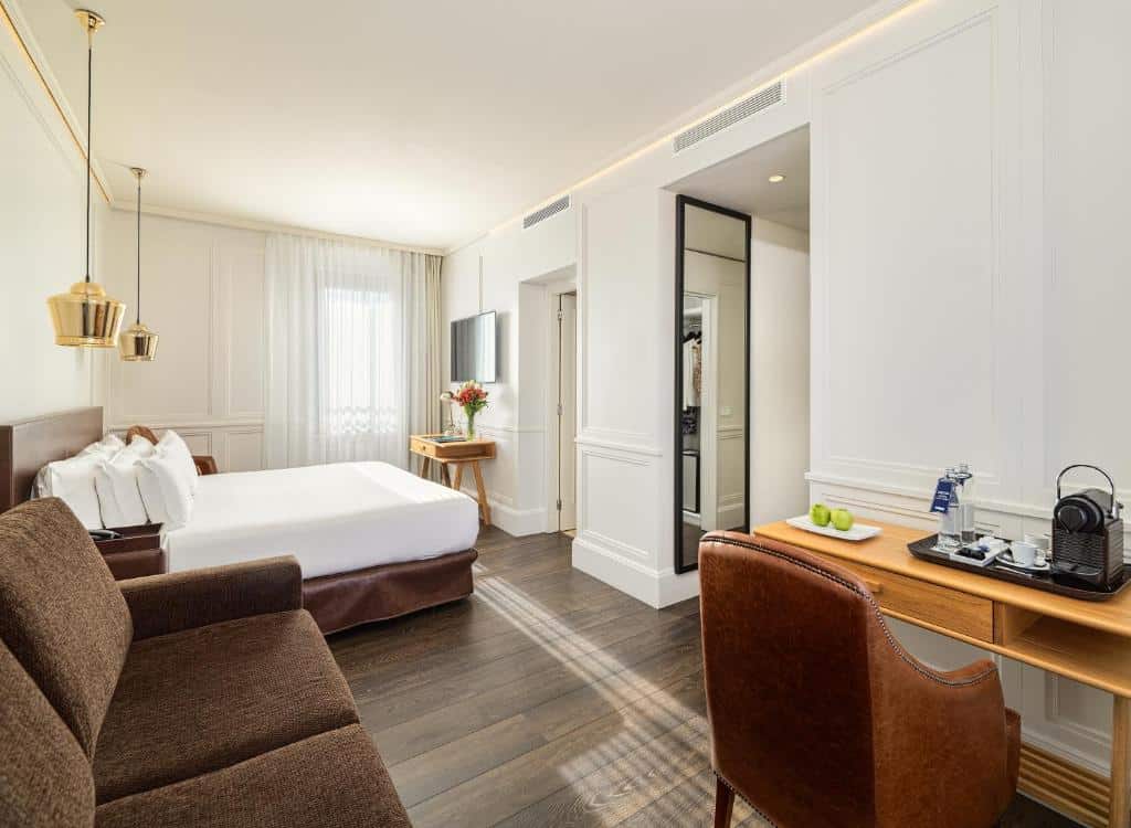 Quarto do Boutique Hotel H10 Montcada. A cama está no fundo do quarto com mesinhas de cabeceira e luminárias dos dois lados. Há uma televisão na parede em frente com uma mesinha abaixo. Na parede ao lado da cama há uma janela.
Um sofá marrom está do outro lado da cama, e uma mesinha com cafeteira e garrafas d'água está na frente junto com uma cadeira.