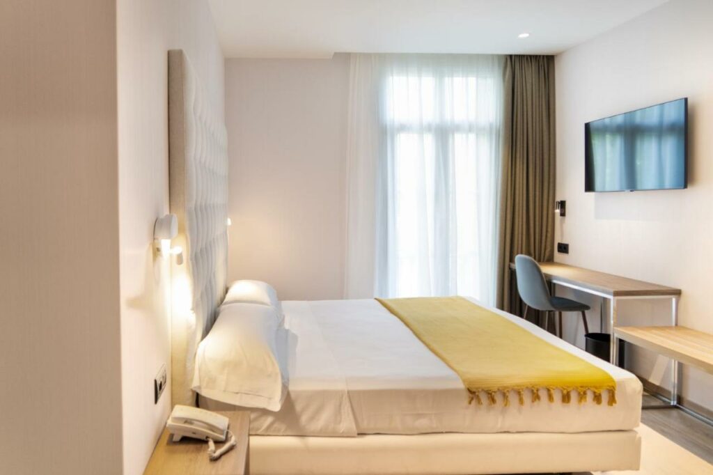 Quarto do hotel Caledonian. Há mesinhas de cabeceira dos dois lados da cama de casal, e uma mesinha de trabalho com cadeira está na frente. Há uma televisão na parede acima da mesinha, e uma janela coberta por cortinas ao lado.