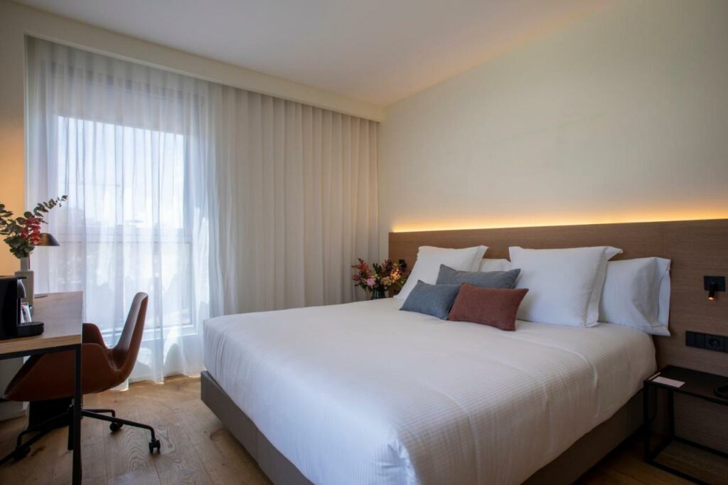 Quarto do Hotel Paxton Barcelona. A cama de casal tem algumas almofadas em cima, e há mesinhas de cabeceira dos dois lados da cama. Uma mesa de trabalho tem cafeteira e um vaso de flores em cima, e há uma cadeira de rodinhas embaixo dela.
Uma janela na parede ao lado está coberta por uma cortina branca.