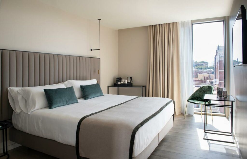 Quarto do Occidental Barcelona 1929, uma das recomendações de hotéis em Barcelona. A cama de lençol e travesseiros verde, bege e brancos está encostada na parede direita e tem um painel acolchoado bege por trás. Há uma luminária pendurada em cima das mesinhas de cabeceira dos dois lados da cama, e uma televisão em frente.
