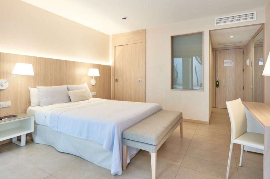 Quarto do Hotel Omnium, uma das recomendações de hotéis em Barcelona. Há uma cama com lençol e travesseiros brancos encostados na parede esquerda na frente de uma porta. Há abajures no painel acima das mesinhas de cabeceira dos dois lados da cama, e um banco aos seus pés.