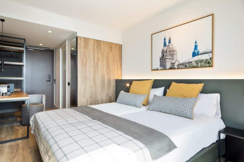 Quarto do Acta Voraport. A cama de casal tem cobertor cinza e almofadas coloridas. Há um quadro na parede acima da cama, e mesinhas de cabeceira dos dois lados. Ao fundo, há uma mesinha com cadeira, uma estante vazia e duas portas.