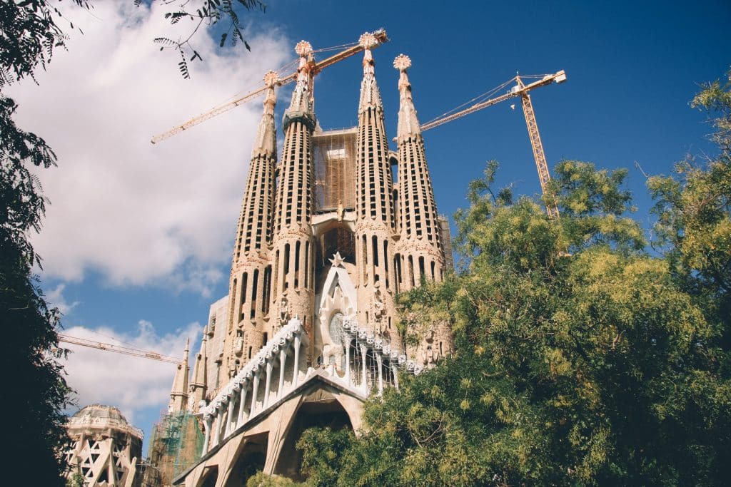 Basílica La Sagrada Familia vista atrás de uma árvore. O céu acima é azul com poucas nuvens. - Foto: iam_os via Unsplash