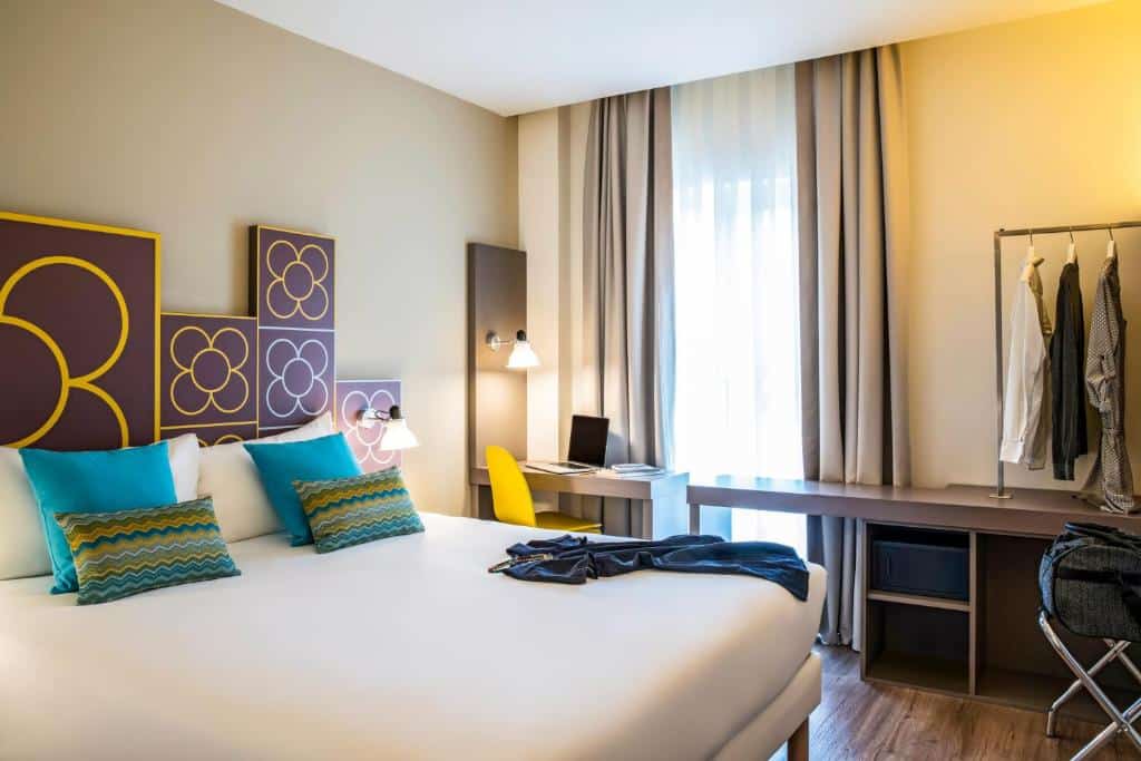 Quarto do ibis Styles Barcelona Centre. A cama de casal tem algumas almofadas azuis e estampadas em cima. Há alguns quadros de flores na parede atrás da cama, e uma mesinha com computador e uma cadeira amarela ao lado da cama. Há uma janela com cortina na frente da mesa, e um suporte de roupas com cofre está do outro lado.