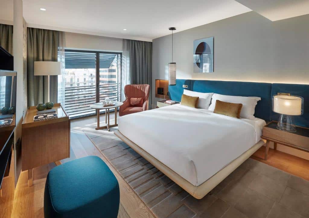 Quarto do Mandarin Oriental Barcelona. A cama está encostada em uma cabeceira acolchoada azul, e há abajures nas mesinhas de cabeceira dos dois lados. Uma televisão está de frente para a cama, com um banquinho azul de um lado e uma cômoda do outro.
Ao fundo há uma janela com cortinas e uma poltrona laranja com uma mesinha á sua frente.