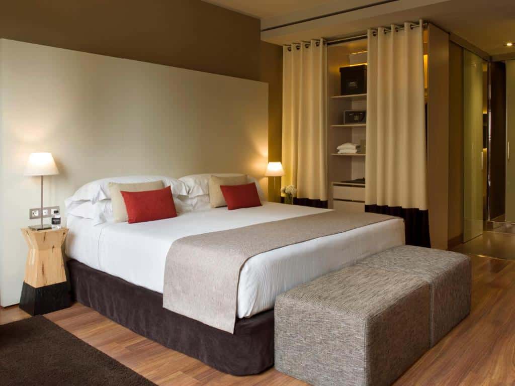 Quarto do Grand Hotel Central. A cama de casal tem algumas almofadas coloridas em cima, e há abajures nas mesinhas de cabeceira dos dos lados da cama. Dois banquinhos estão aos pés da cama. Há um armário com cortinas ao lado.
