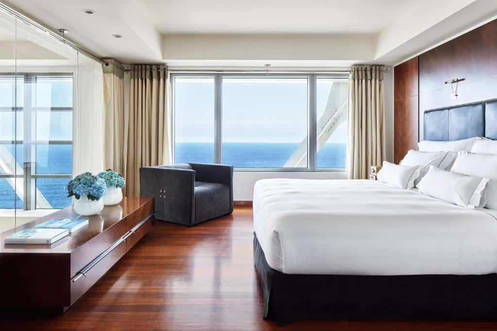 Quarto do Hotel Arts Barcelona. A cama de casal está encostada na parede direita, e há uma cômoda com plantas e livros em cima à sua frente. Há um espelho em cima da ômoda. Ao fundo, uma janela com vista para o mar tem uma poltrona marrom ao lado.