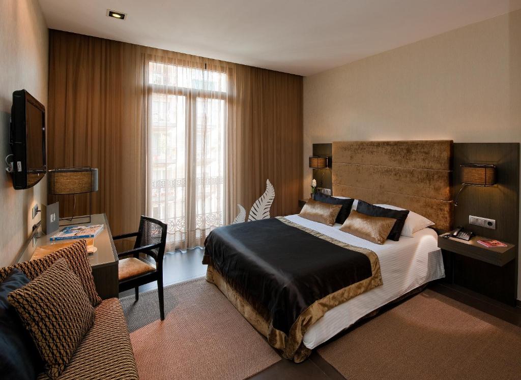 Quarto do Hotel Constanza. Há uma cama encostada na parede com lençol e travesseiros marrom e pretos. Há um painel marrom atrás da cama, e abajures acima das mesinhas de cabeceira dos dois lados.
Uma mesa de trabalho com cadeira estão de frente para a cama. Uma televisão está na parede acima da mesa, e há um abajur de um lado e uma poltrona do outro. Há dois tapetes no quarto e uma escultura de folha branca perto da janela com cortinhas marrons fechadas.