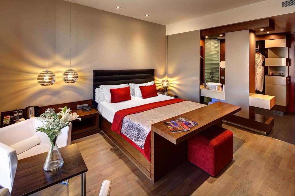 Quarto do Hotel Olivia Plaza Hotel, uma das recomendações de hotéis em Barcelona. A cama tem lençol e travesseiros brancos e vermelhos. Há uma mesinha de madeira aos pés da cama com revistas em cima e um pufe vermelho abaixo. Há abajures acima das mesinhas de cabeceira dos dois lados da cama.