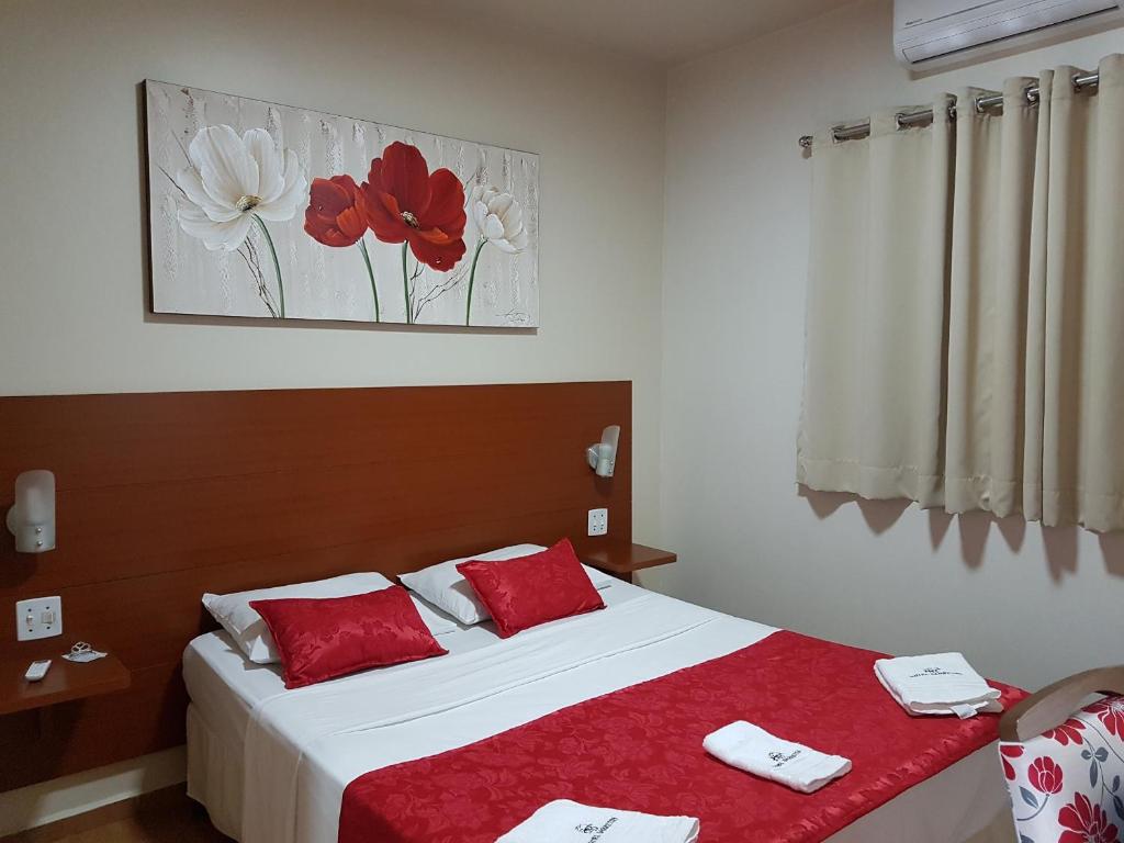 Uma cama bem arrumada com detalhes em vermelho, uma pintura floral acima, cortinas bege e toalhas colocadas sobre a cama, em um dos quartos do Hotel Barretos.