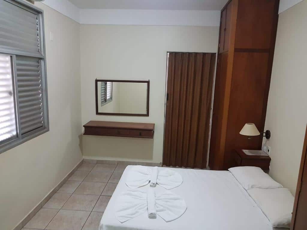 Quarto com cama de casal, um abajur do lado, móveis de madeira, janela e um espelho na parede branca, no Scala Hotel Barretos, para representar hotéis em Barretos.
